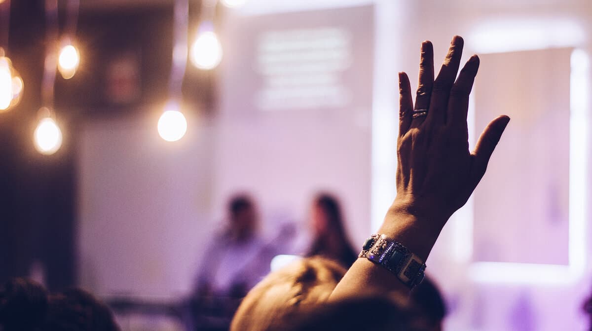 A stage during a seminar in the background with a raised hand in the foreground